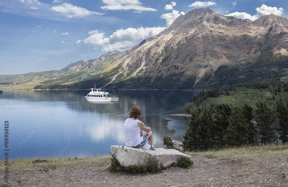 horizontal image of a lady sitting on a boulder and watching a boat glide by on the clear lake flanked by mountains.