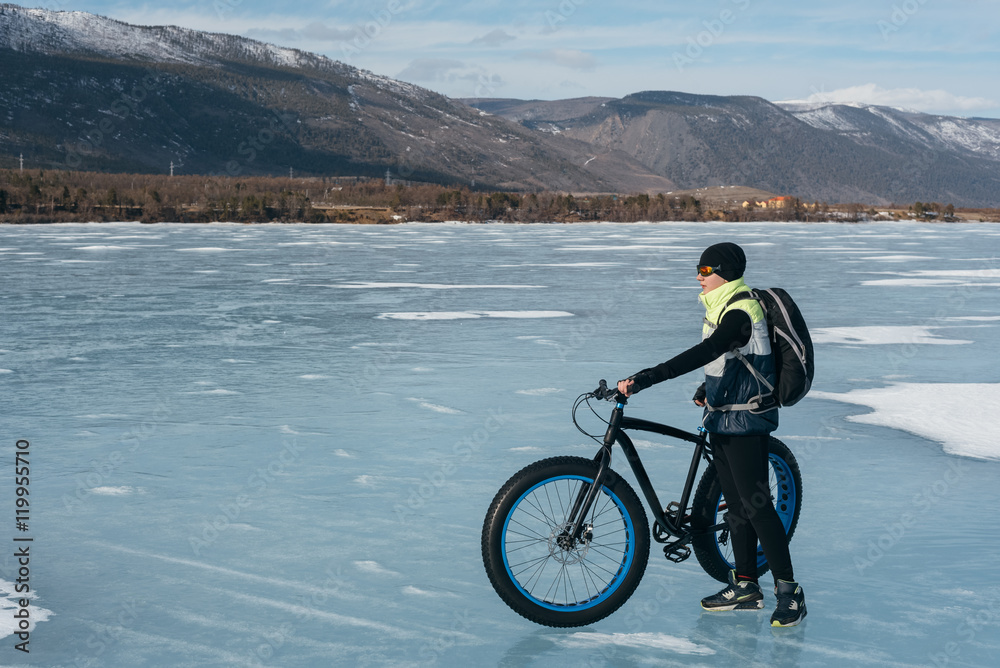 Obraz premium Fatbike (also called fat bike or fat-tire bike) - Cycling on large wheels. Cyclist holding a bike, and watching the sunset. They are standing on the frozen lake.
