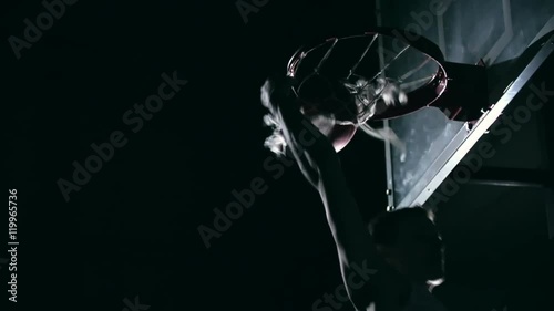 Low angle of slam dunk performing by muscular basketball player in dark court in slow motion