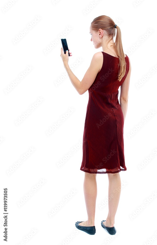 back view of standing young beautiful woman using a mobile phone. girl watching. Isolated over white background. The girl in the maroon sleeveless dress looks into the smartphone. back view of standing young beautiful woman using a mobile phone. girl watching. Isolated over white background. The girl in the maroon sleeveless dress looks into the smartphone.