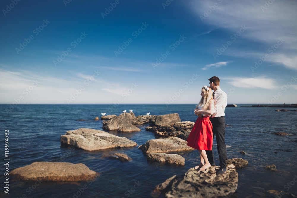 Romantic loving couple posing on stones near sea, blue sky