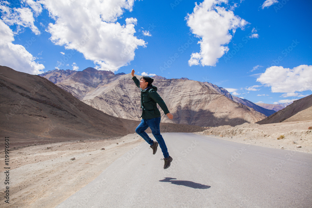 Extremely happy young male tourist jumping on country road with giant mountains behind