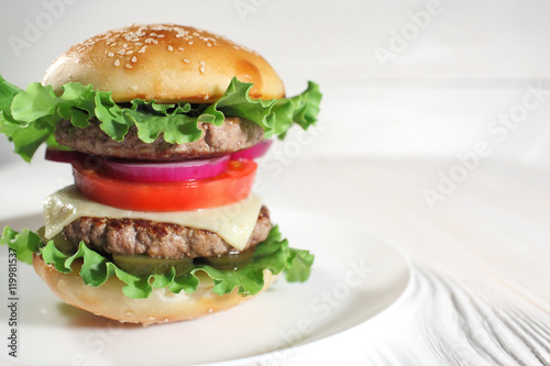 Big juicy traditional American burger with grilled meat and slices fresh vegetables closeup on a white plate on a wooden table. Food and beverages. Selective focus with shallow depth of field
