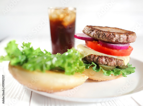 Big juicy traditional American burger with grilled meat and slices fresh vegetables closeup on a white plate on a wooden table. Food and beverages. Selective focus with shallow depth of field