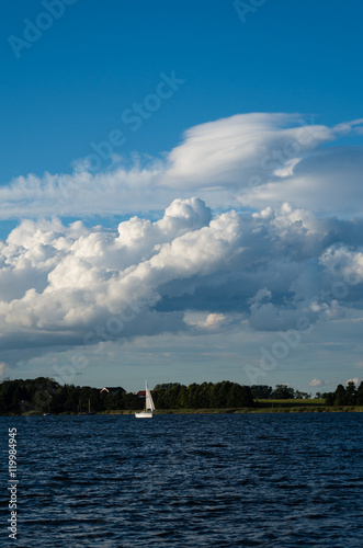 Widok na jezioro w po którym płynie jacht żaglowy. Na niebie widać rozbudowujące się chmury Cumulus.