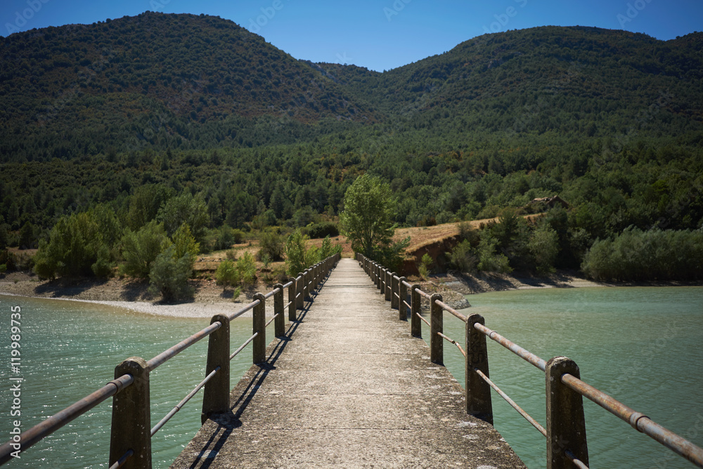 Naklejka premium Walking on a concrete pedestrian bridge over a shallow light blue bay towards wooded hills