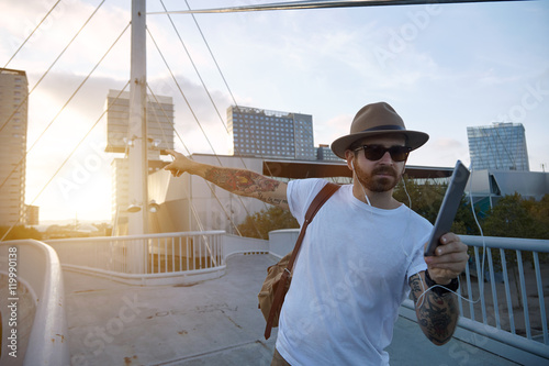 A young traveler wearing a hat, sunglasses and an unlabeled white t-shirt points to something during a video call on a bridge in a city at sunset