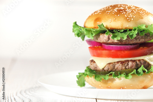 Big juicy traditional American burger with grilled meat and slices fresh vegetables closeup on a white plate on a wooden table. Food and beverages. Selective focus with shallow depth of field