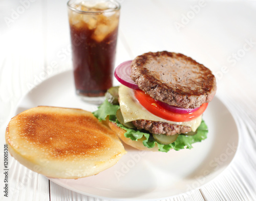 Big juicy traditional American burger with grilled meat and slices fresh vegetables closeup on a white plate on a wooden table. Food and beverages. Selective focus with shallow depth of field