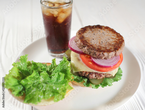 Big juicy traditional American burger with grilled meat and slices fresh vegetables closeup on a white plate on a wooden table. Food and beverages. Selective focus with shallow depth of field