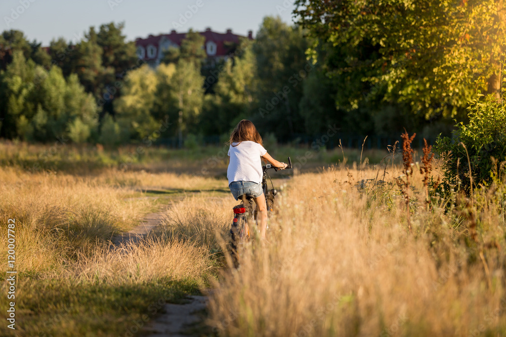 Woman riding bicycle at meadow on dirt road at sunset