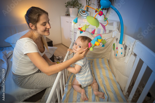 Portrait of young mother looking at her baby in crib before goin photo