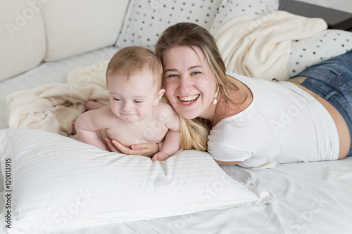 Cheerful laughing mother relaxing on bed with her 9 months old b