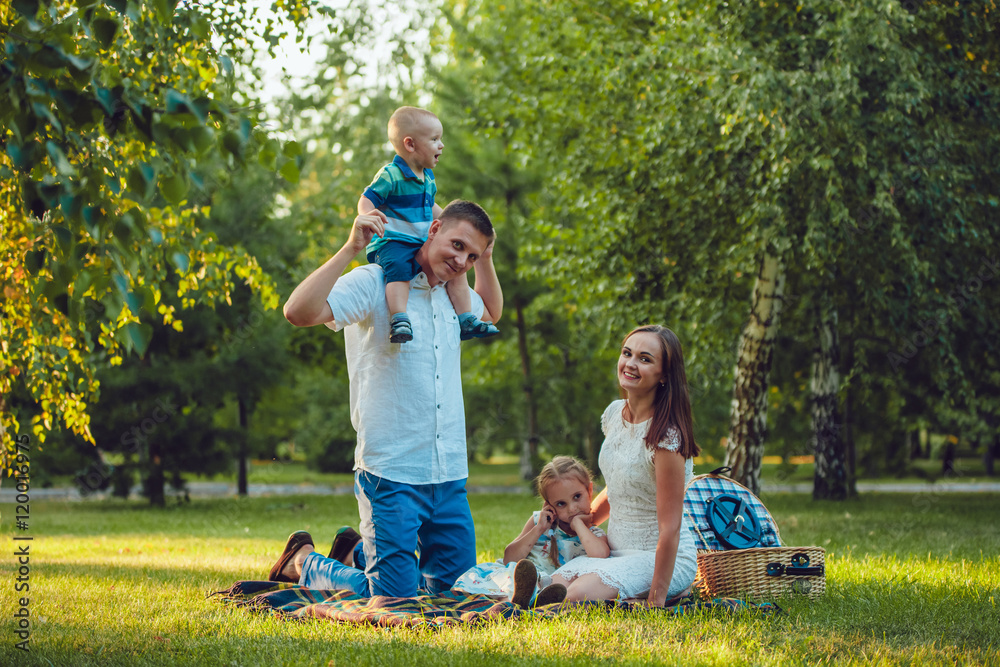 Fototapeta premium Young happy family of four on picnic in the park and father holding his son shoulders