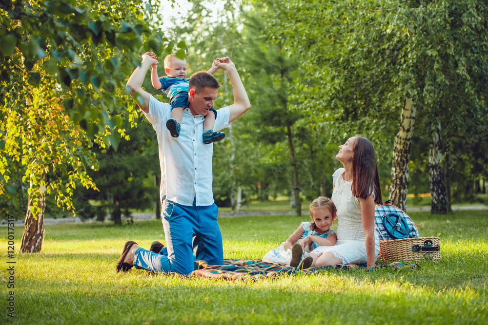 Fototapeta premium Young happy family of four on picnic in the park and father holding his son shoulders