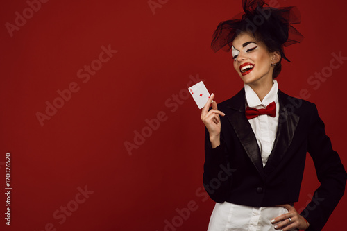 Bilde på lerret Portrait of a young beautiful lady croupier with an artistic make up joker on the red background holding an ace card and laughing