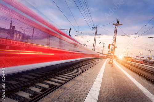 High speed red passenger train on railroad track in motion at beautiful sunset. Blurred commuter train. Railway station in the evening. Railroad travel, railway tourism. Industrial landscape. Train
