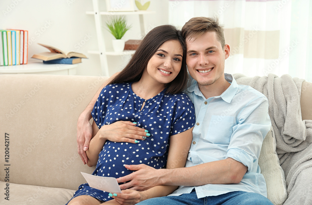 Beautiful young pregnant couple with ultrasound image sitting on sofa