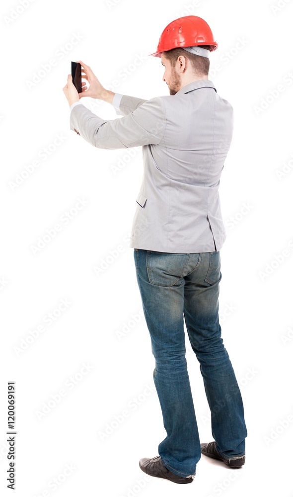 Backview of business man in construction helmet stands and enjoys tablet or using a mobile phone. Standing young guy. guy in a gray jacket and an orange helmet takes on the mobile phone.... Backview of business man in construction helmet stands and enjoys tablet or using a mobile phone. Standing young guy. guy in a gray jacket and an orange helmet takes on the mobile phone....
