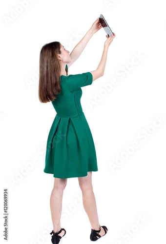 back view of standing young beautiful woman using a mobile phone. girl watching. backside view of person. slender brunette in a green short dress takes on something at the top of the plate. © ghoststone