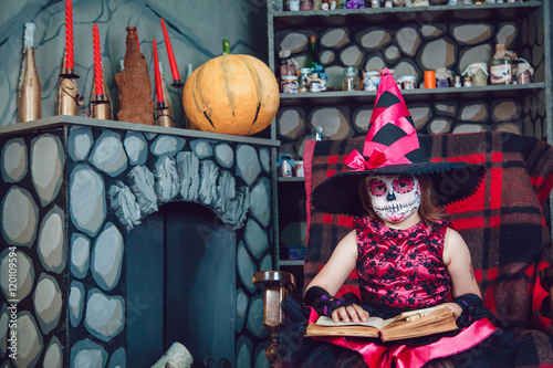 Girl in witch costume and make-up on her face sitting in a chair with book in his hands in halloween decorations