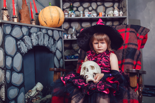 Girl in a witch costume sitting in chair with skull in his hands in in halloween decorations