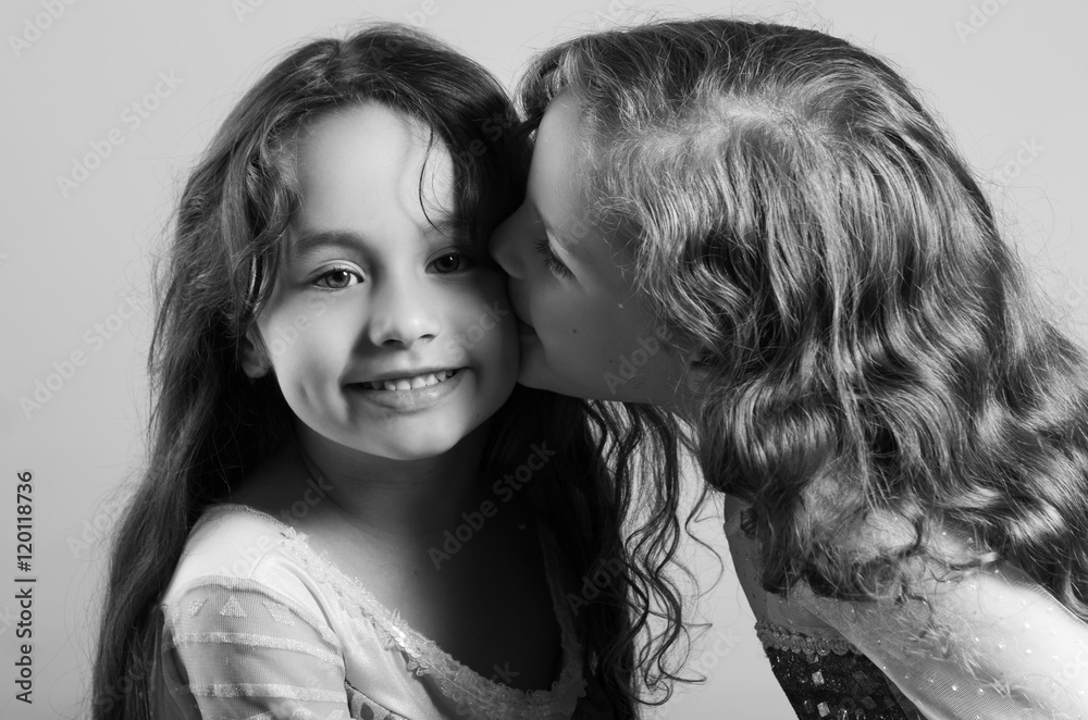 Adorable big sister kissing sibling on cheek, both wearing matcing blue dresses posing together happily, studio background Adorable big sister kissing sibling on cheek, both wearing matcing blue dresses posing together happily, studio background