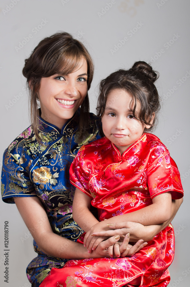 Mother with little daughter dressed in beautiful chinese blue and red dresses posing happily embracing for camera, studio background Mother with little daughter dressed in beautiful chinese blue and red dresses posing happily embracing for camera, studio background