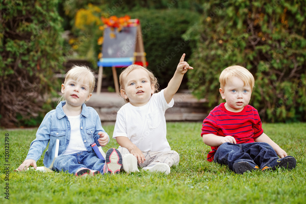 Group of three white Caucasian toddler children kids boys and girl sitting outside in summer autumn park by drawing easel with fall leaves looking in camera, back to school Group of three white Caucasian toddler children kids boys and girl sitting outside in summer autumn park by drawing easel with fall leaves looking in camera, back to school