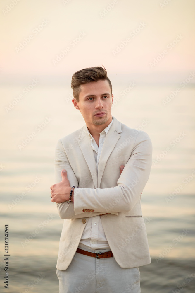 young handsome man in a white suit near the sea at sunset. young handsome groom on the beach. Close-up young handsome man in a white suit near the sea at sunset. young handsome groom on the beach. Close-up