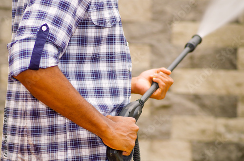 Closeup arms of man wearing square pattern blue and white shirt holding high pressure water gun, pointing towards grey brick wall photo