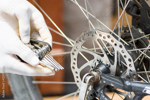 Closeup hand wearing white glove holding multi unbrako key tool next to parts of bicycle wheel, mechanical repair concept