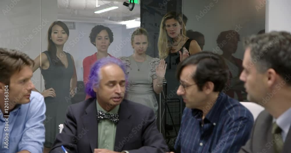 Business men have a meeting while female colleagues are left standing outside glass walls of conference room. Medium shot, low angle, recorded hand held in real time with focus pull. Business men have a meeting while female colleagues are left standing outside glass walls of conference room. Medium shot, low angle, recorded hand held in real time with focus pull.