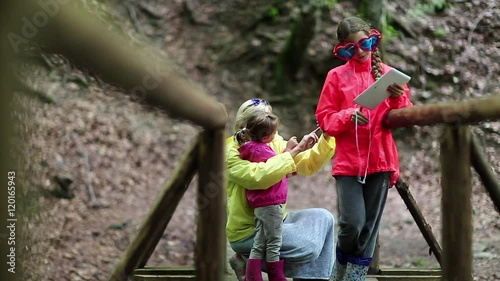 Mother and two daughters with smartphone and tablet computer on the nature. Family with electronic devices on the wooden bridge in the forest. Women in color windcheaters with phone and tablet pc photo