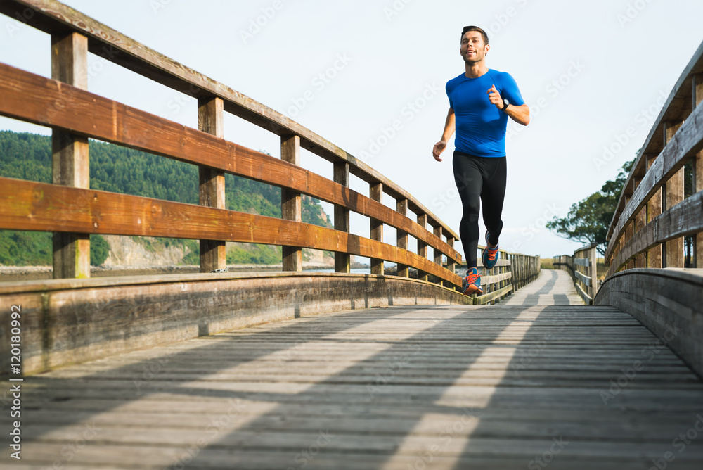 Smiling man running in park on wooden walkway training and exercising for trail run marathon race. Fitness healthy lifestyle concept with male athlete outdoor runner.