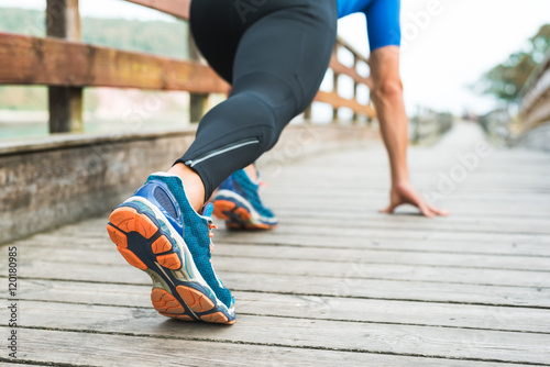 Fitness man ready for running on wooden walkway on an outdoor park. Male athlete in powerful confident starting line pose in Rodiles, Asturias. Sport Shoe sole closeup. photo