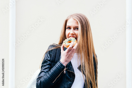 Beautiful girl bites a delicious donut walking on the streets of European city. Warm spring. Outdoor. Close up