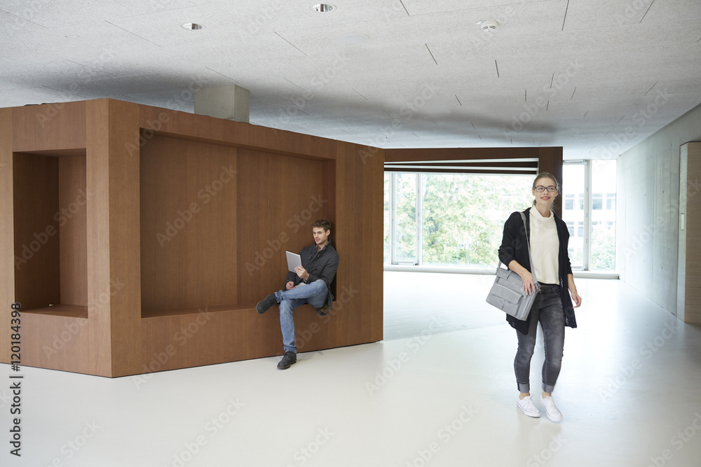 Young man sitting on wooden ledge using tablet with woman passing by Young man sitting on wooden ledge using tablet with woman passing by