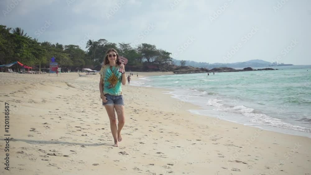 Young Long Haired Hippie Girl Walks along the Beach at a Sunny Day Holding an Ukulele in Her Hand. Slow Motion Young Long Haired Hippie Girl Walks along the Beach at a Sunny Day Holding an Ukulele in Her Hand. Slow Motion