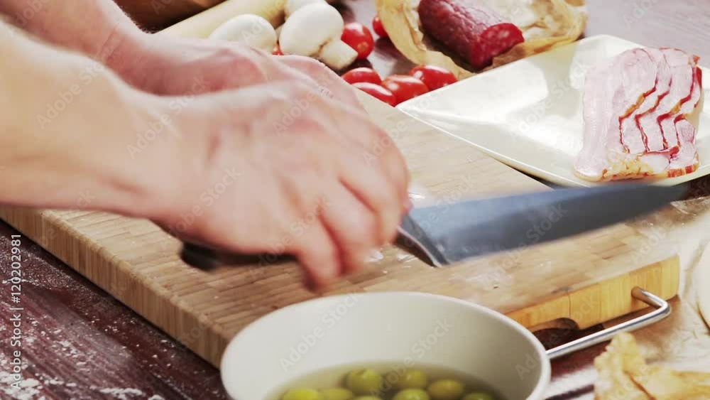 Mens hands cut olives on bamboo cutting board for cooking pizza. Pizza ingredients on wooden background Mens hands cut olives on bamboo cutting board for cooking pizza. Pizza ingredients on wooden background