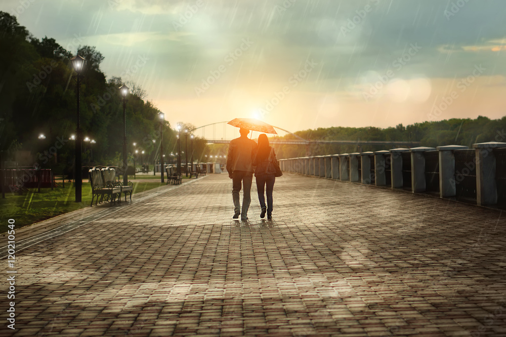 Naklejka premium Widok para pod parasolem idąc w dół deszczowego parku w jesienny wieczór. Jasny zachód słońca.