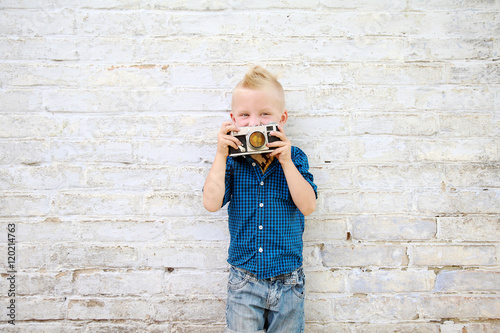 мальчик с фотоаппаратом фотографирует возле белой кирпичной стены 