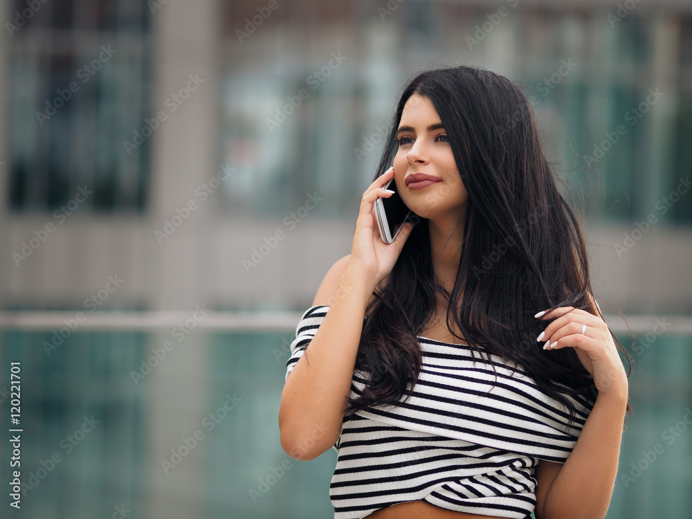 Portrait of young beautiful curvy woman plus size in striped top and black skirt over blurred city background using her white smart phone and talking Portrait of young beautiful curvy woman plus size in striped top and black skirt over blurred city background using her white smart phone and talking