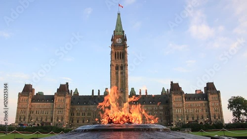 Centennial Flame burning brightly in front of Peace Tower and Centre Block on Parliament Hill, Ottawa, Canada. Parliament Building was lit by morning sun light under blue sky and white clouds