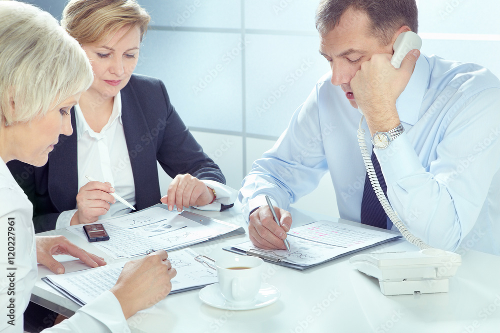 Obraz premium Businessman talking on telephone and writing on paper with two businesswomen working near by