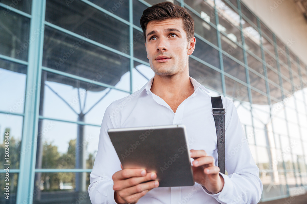 Pensive handsome young businessman standing and using tablet