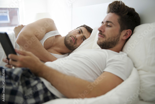 Gay Male Couple In Bed At Home Looking At Digital Tablet