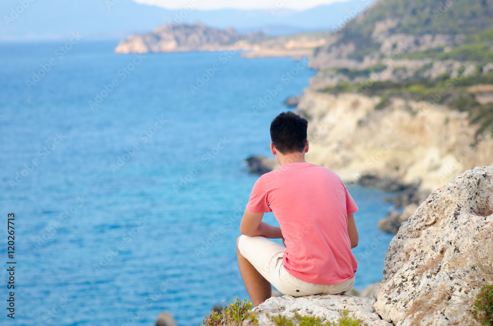 Teenager man sit melancholic on rock on beach near deep blue sea