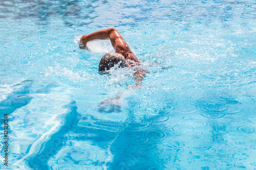 Senior man swimming front crawl  freestyle in an outside pool on a summer  sunny day. The pool steps can be seen and the splashes from the swim.