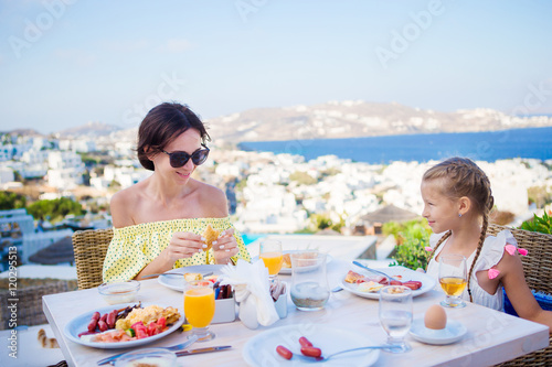 Obraz na płótnie Family having breakfast at outdoor cafe with amazing view on Mykonos town. Adorable girl and mom drinking fresh juice and eating croissant on luxury hotel terrace w salonie